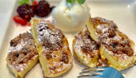 Auf einem weißen Teller liegen vier goldbraune, mit Puderzucker bestreute Ananas-Küchlein. Dazu gibt es eine weiße Eiskugel, die von glänzenden Beeren und einem Minzblatt umgeben ist. Eine silberne Gabel liegt schon bereit, um dieses süße Dessert zu kosten.