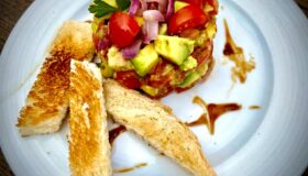Avocado-Tomaten-Tatar auf einem weißen Teller angerichtet und ein paar Streifen Toastbrot dazu.