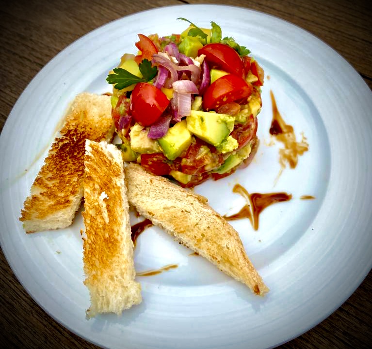 Avocado-Tomaten-Tatar auf einem weißen Teller angerichtet und ein paar Streifen Toastbrot dazu.