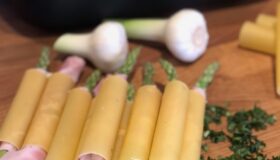Grüner Spargel mit Schinken umwickelt in Cannelloni-Pasta auf einem Holzbrett neben einer dunklen Auflaufform und frischen Kräutern.
