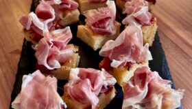 Focaccia in Würfel geschnitten mit einer kurz angebratenen Tomate und Parmaschinken garniert und auf einem schwarzen Teller angerichtet.