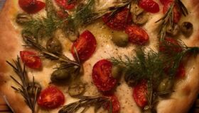 Ein fertiges Focaccia mit Tomaten, Rosmarien und Oliven auf einem Hozbrett.