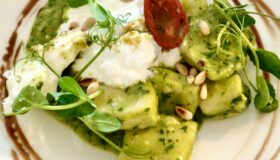 Kartoffelgnocchi mit grünem Pesto, Burrata, Pinienkerne und Kresse auf einem weißen Teller angerichtet.