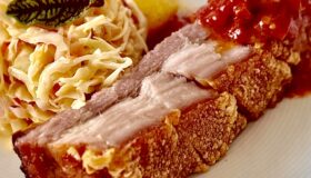 Eine Scheibe Krustenbraten mit Krautsalat und Kartoffeln auf einem weißen Teller.