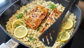 Gebratenes Lachsfilet auf Orzo-Pasta (Kritharaki) mit Zitronenscheiben und frischen Kräutern in einer quadratischen, schwarzen Pfanne auf einem Induktionsherd.