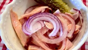 Leberkäs-Wurstsalt in einer kleinen weißen Schüssel mit einer sauren Gurke und roten Zwiebeln angerichtet.
