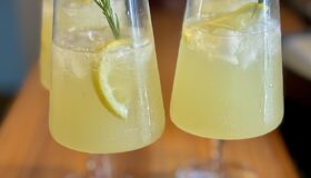 Limoncello Spritz in vier Gläsern angerichtet und mit Zitrone und einem Zweig Rosmarien garniert.