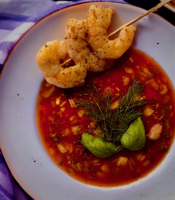 Eine kalte Melonensuppe mit drei knusprigen Garnelen am Spieß auf einem Teller serviert und mit Basilikum garniert.