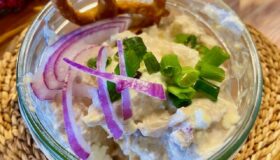Obatzter in einem Glas angerichtet und mit Zwiebeln und Schnittlauch und einer kleinen Bezen garniert.