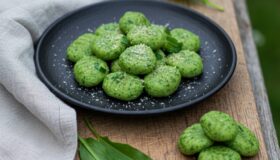 Grüne Bärlauch-Gnocchi mit Parmesan bestreut auf einem schwarzen Teller, serviert auf einem Holzbrett neben frischen Bärlauchblättern.