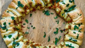 Pizza als Kranz geformt mit frischem grünem Pesto und Cheddar-Käse gefüllt auf einer grauen Schieferplatte dekoriert und mit Kräutern oben drauf.