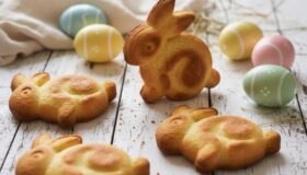 Vier goldbraun gebackene Osterhasen aus Rührteig auf einem rustikalen weißen Holztisch, dekoriert mit bunten Ostereiern und Leinentuch.