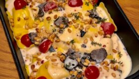 Gefüllte Ravioli in cremiger Sauce mit schwarzen Trüffelscheiben, Kirschtomaten und gerösteten Pinienkernen in einer rechteckigen schwarzen Auflaufform von Pfannen Harecker.