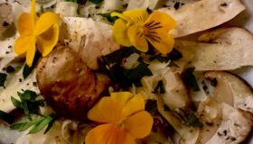 Auf einem Tisch liegt ein weißer Teller mit Steinpilz Carpaccio, dekoriert mit essbaren Blumen und Parmesan.