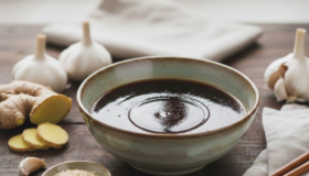Selbstgemachte Teriyake Sauce in einer grauen Schale. Im Bild sind Ingwer, Knoblauch und Sesamkörner enthalten, sowie 2 Eßstäbchen