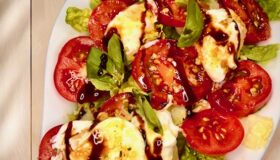 Tomatensalat mit Burrata und frischem Basilikum in einer Öl-Balsamico-Sauce auf weißem Teller angerichtet.