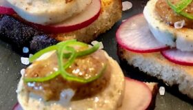 Drei Weißwurst Canapès mit Radieschen und süßen Senf garniert auf einem schwarzen Teller mit Salz angerichtet.