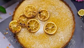 Zitronen-Tarte, ein frisches und saftiges Rezept. Tarte platziert auf einer grauen Platte mit gebackenen Zitronen oben drauf, Blumen dazu für die Deko.