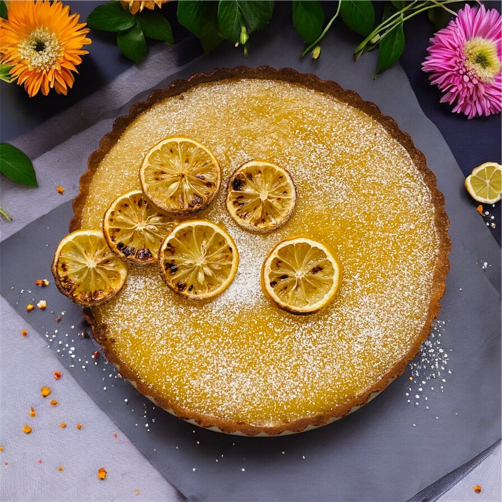 Zitronen-Tarte, ein frisches und saftiges Rezept. Tarte platziert auf einer grauen Platte mit gebackenen Zitronen oben drauf, Blumen dazu für die Deko.
